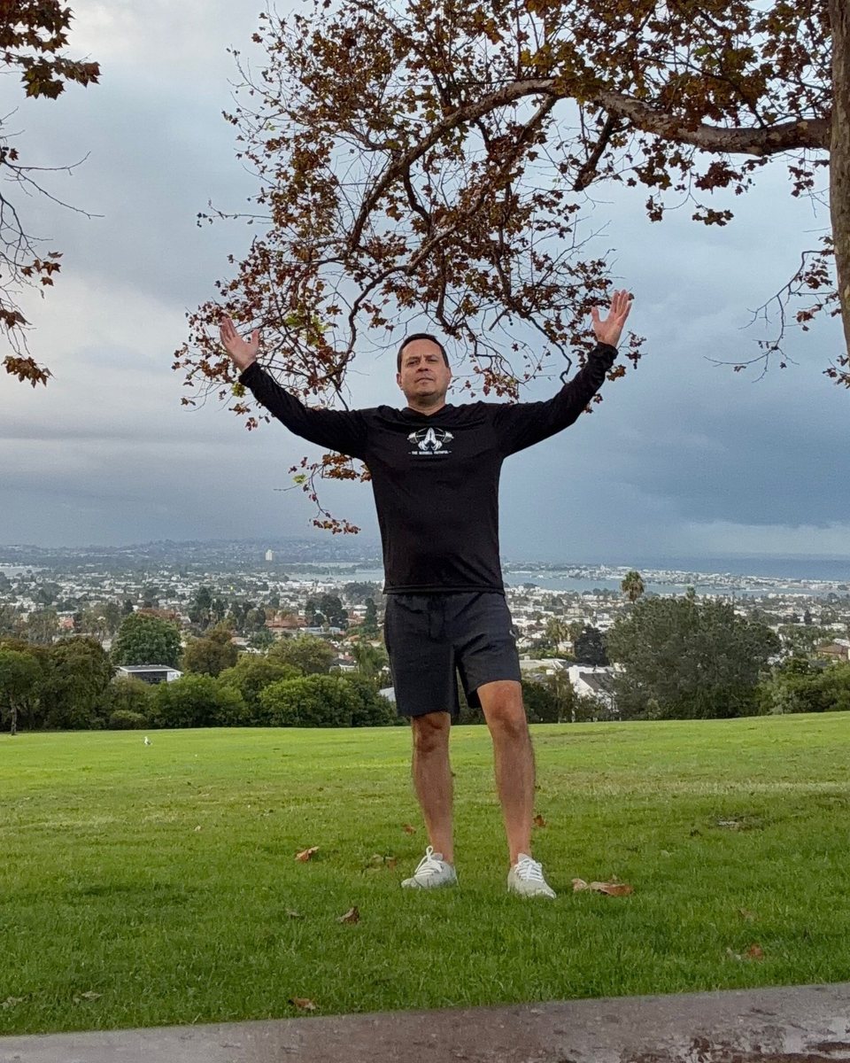 Jeremy with arms raised on a hilltop in San Diego, wearing the black Barbell Faithful performance hoodie
